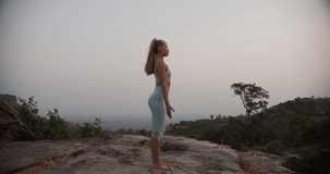 Woman standing straight barefoot on the rocks as she stretches both her hands and ending the clip doing the prayer hands and a beautiful scenic view of the mountains in the background.
