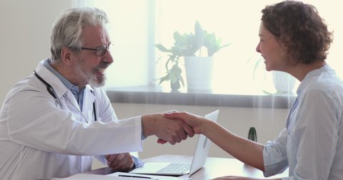 Side view smiling young female patient shaking hands with happy middle aged mature male doctor in white uniform, thanking for help or feeling excited about healthcare treatment results at meeting.