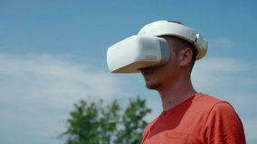 Man in a virtual reality helmet turns head down and to the right. The guy in the park in a red T-shirt on a background of blue sky and green trees looks VR glasses. 4K footage
