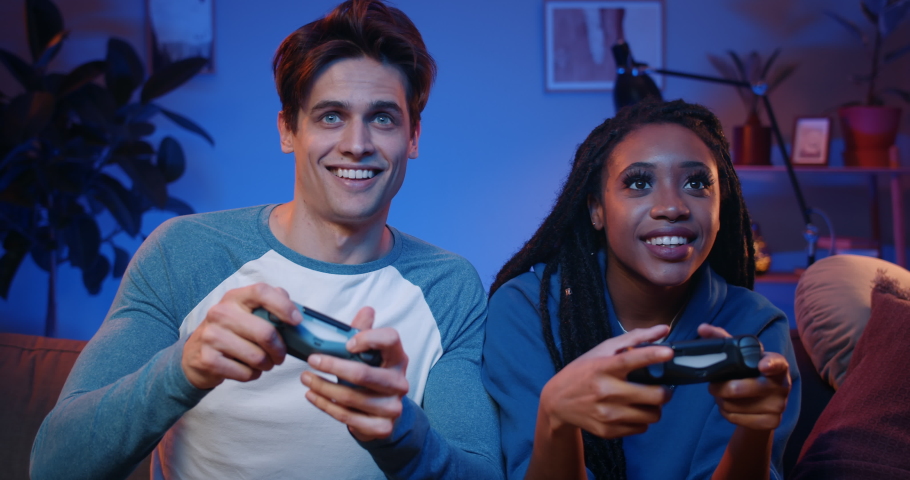 Happy young people playing video games on console while sitting on couch in front of tv. Millennial couple spending fun time together at home.Room with warm and neon lights. | Shutterstock HD Video #1054788125
