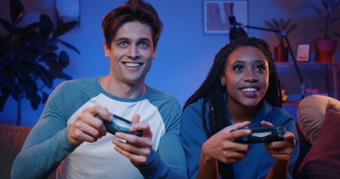Happy young people playing video games on console while sitting on couch in front of tv. Millennial couple spending fun time together at home.Room with warm and neon lights.
