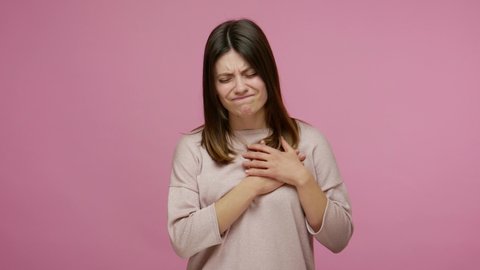 Depressed worried young woman suffering intense pain in chest, frowning from heartache, painful cramps of cardiac problems, risk of infarction, stroke. indoor studio shot isolated on pink background