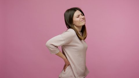 Upset overworked woman massaging sore spine, stiff muscles, suffering backache, painful spasms from sedentary lifestyle, risk of rheumatoid arthritis. indoor studio shot isolated on pink background