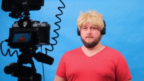 freaky fat man in a wig and a pink t-shirt makes a video blog against a blue background. High quality 4k footage