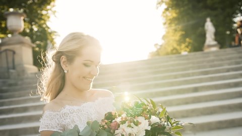 Happy bride walks in a park in Paris. Beautiful blonde descends the stairs at sunset in a white dress and with a bouquet of flowers