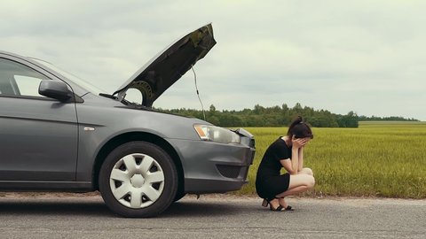 An upset girl in a short dress sits on the pavement and waits for help after her car breaks down on the road. Slow motion.