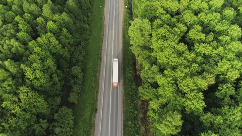 One Semi Truck with white trailer and cab driving / traveling alone on dense flat forest asphalt straight empty road, highway top down view follow vehicle aerial footage / Freeway trucks traffic