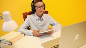Happy teen guy in headphones using a laptop computer, learning through an online e-learning system, on a yellow background