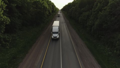One Semi Truck with white trailer and cab bonnet driving / traveling alone on dense flat forest asphalt straight empty road, highway wide view follow vehicle aerial footage / Freeway trucks traffic