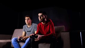 Having fun enjoy victory. Emotional two young guys are sitting on the couch in front of the TV and excited playing a game console, holding a joystick. Mens gaming with a wireless controller. Cozy room