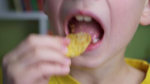 Smiling boy enjoys chips. Young caucasian kid eating unhealthy potatoes crisps. Child puts potato chips in his mouth. Snack on junk food. Portrait of a boy with a satisfied face. Tasty food.