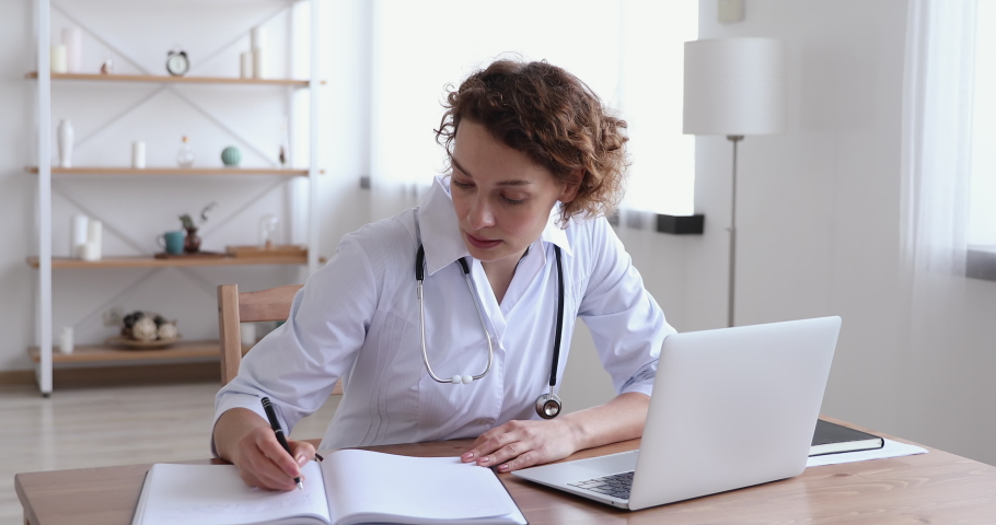 Focused 30s female general practitioner in white uniform sitting at workplace, doing paperwork and using medical software application on computer, involved in research or making report at clinic. Royalty-Free Stock Footage #1054813136