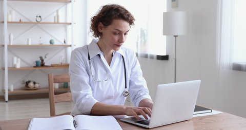 Focused 30s female general practitioner in white uniform sitting at workplace, doing paperwork and using medical software application on computer, involved in research or making report at clinic.