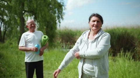 elderly woman leads sporty lifestyle and performs exercises for hands from joint pain background of husband during workout outdoors on warm sunny day by river