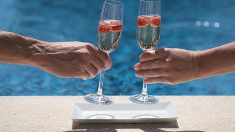 Close up of glasses with champagne or prosecco with raspberry on swimming pool or sea background. Happy white woman and man take and clink glasses. Romantic weekend, vacation for two, honeymoon. 