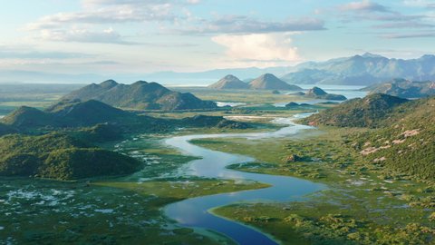 Blue river flowing through green valley toward distant mountains. Bends and curves of Crnojevica river in Montenegro, meandering through the marsh among hills on its way to lake Skadar. Aerial footage