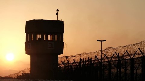 A silhouette of a prison watchtower at sunset, the barbed wire is apparent at the bottom of the frame and the sun ball itself as well.