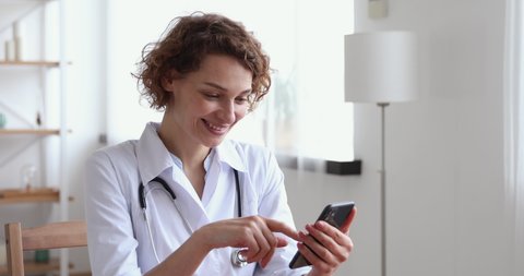 Head shot smiling young female physician in white medical coat sitting at desk, using mobile healthcare application, consulting patients online. Happy medical worker checking messages in smart phone.