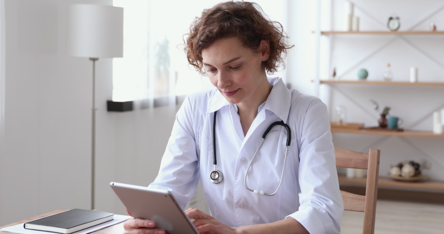 Happy young female general practitioner reading email with good news from patient or enjoying working online on digital tablet. Smiling trustful 30s therapist consulting client distantly at workplace. | Shutterstock HD Video #1054818062