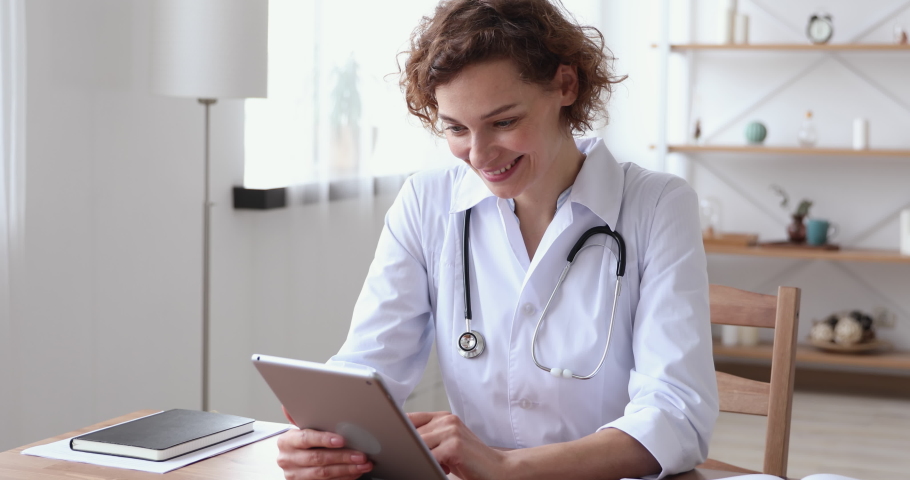 Happy young female general practitioner reading email with good news from patient or enjoying working online on digital tablet. Smiling trustful 30s therapist consulting client distantly at workplace. Royalty-Free Stock Footage #1054818062