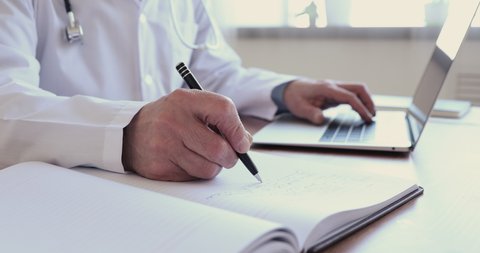 Close up wrinkled male hands writing information in medical registration journal. Old mature doctor therapist working at office, using healthcare software application on computer, handwriting notes.