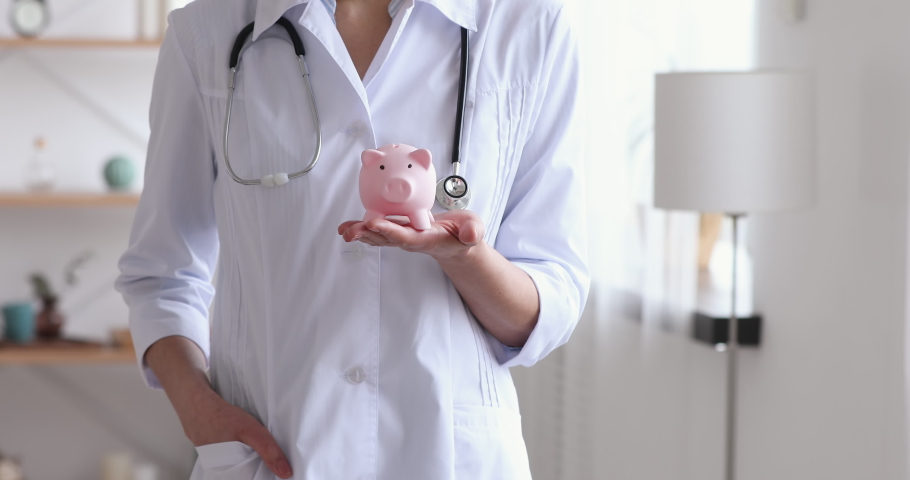 Smiling young female general practitioner in white coat holding piggy bank box in hands, money savings for health insurance, medical services investment, medicine cost, charity aid donation concept. Royalty-Free Stock Footage #1054819934