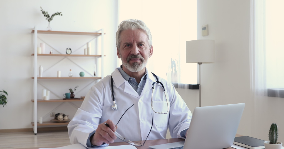 Portrait of confident old mature male head doctor physician in white medical uniform holding glasses sitting at workplace with computer. Happy general practitioner looking at camera, posing in office. Royalty-Free Stock Footage #1054822709