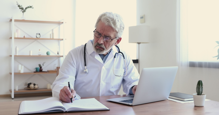 Focused middle aged senior head doctor in white medical coat and glasses sitting at workplace, web surfing information online or using healthcare computer application, writing notes in paper journal. Royalty-Free Stock Footage #1054822712