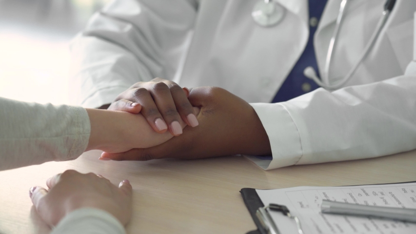 African female doctor holding hand supporting caucasian woman patient. Kind ethnic professional physician give empathy concept encourage reassure infertile patient at medical visit, close up view. | Shutterstock HD Video #1054823156