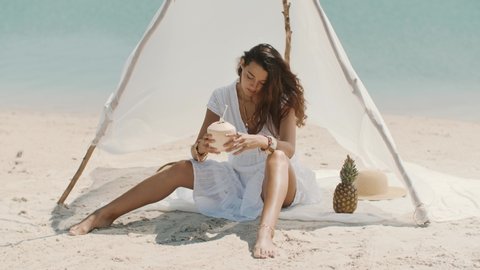 Woman drinking coconut juice while relaxing on the beach near the water. Tropical fruits diet concept. Summer holiday idyllic. Happy young slim mixed race Asian Caucasian girl in bikini and hat