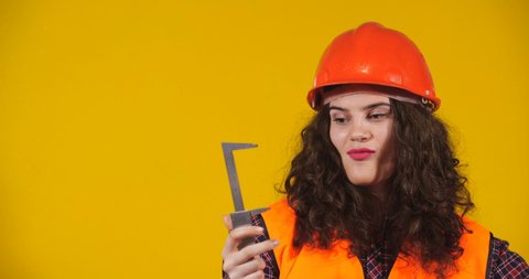 Attractive plus-size model posing. Curly-haired brunette on bright yellow studio background. Girl in red helmet and orange signal vest showing large size with caliper. Erotic Fantasy Concept