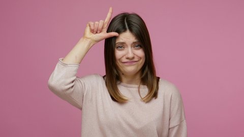 You lose! Brunette young woman making loser gesture, L sign on forehead and pointing to camera, teasing and accusing for defeat, expressing disrespect. indoor studio shot isolated on pink background