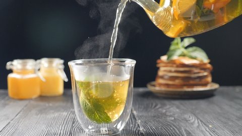 Pouring tea with orange, lemon and lime out of transparent tea pot into glass with nint.Brewing tea with honey and pancakes in background. Slow motion tea. Full hd