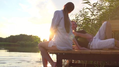 Beautiful couple relaxing and drinking red wine on the Pier over sunset. People Enjoying nature outdoor near river. Love, vacation concept. Bare feet in a water. Freedom, happiness. Slow motion 4K UHD