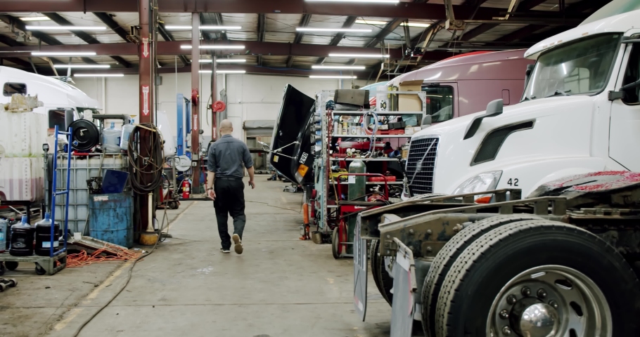 The master in the repair service of trucks goes from the camera to the depths of the shop.