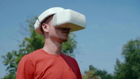 Man in a virtual reality helmet is studying virtual game. The guy in the park in a red T-shirt on a background of blue sky and green trees looks VR glasses. 4K footage