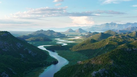River meandering through the valley toward distant mountains, in the morning. Crnojevica river in Montenegro, flowing through the marsh, among hills, on its way to lake Skadar. Aerial footage.