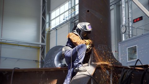 Professional heavy industry worker welder in protective mask and suit works with metal construction on factory. Steel and iron welding construction site. Welding machine with sparks and flashes.