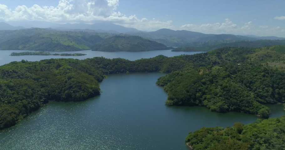 Gorgeous blue water lakes surrounded by hills with lush green trees and foliage with Cordillera Central (Hispaniola) mountain range in background, overhead rising aerial Royalty-Free Stock Footage #1054847816