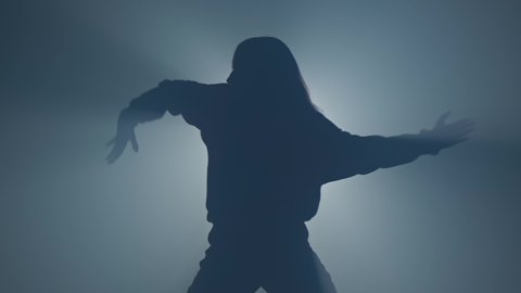 Silhouette of woman in hoody performing hip hop moves. Graceful female dancing in studio with smoke and spotlight. 4K, UHD