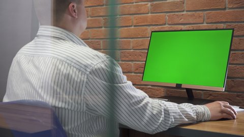 Young Man Working On Computer With Green Screen In Office. Man Works On  Personal Computer With Green Mock-Up Screen In  Office. Man Sits At  Table In Office And Works On Computer. Green Screen