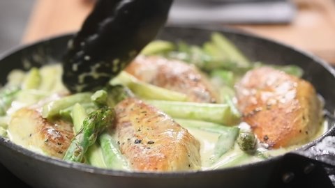 Chef preparing delicious chicken breast with asparagus in a creamy sauce in frying pan.