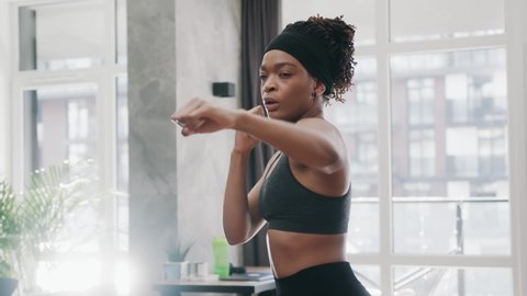 Serious african amerian athletic woman punching the air with her fists training warm-up before boxing workout at home interior. Sports. Activity. Motivation. Confidence.