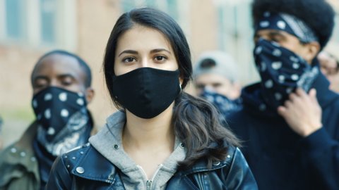 Close up of young Caucasian beautiful woman in mask looking straight at camera outdoor at street riot. Portrait of girl with male Afrcan Americans on background. Female protester at manifestation.