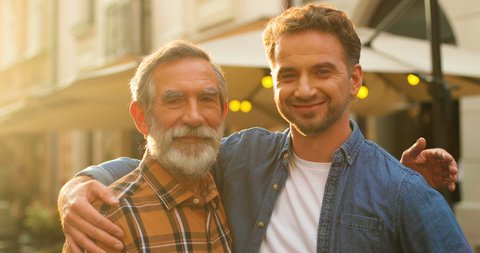 Portrait of happy Caucasian male generations at street posing to camera. Young handsome adult man with senior grandfather. Old gray-haired dad with son smiling and embracing outdoors.