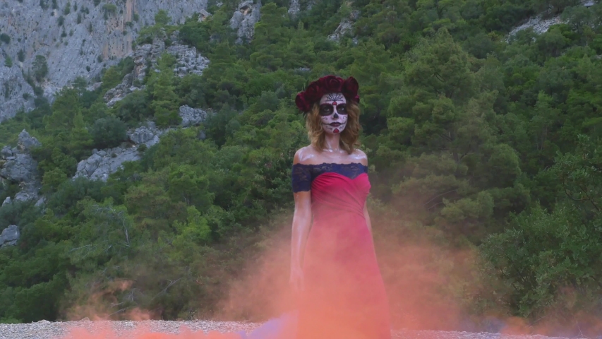 Santa Muerte girl in pink dress with bouquet on Halloween eve outdoors in red and blue smoke
