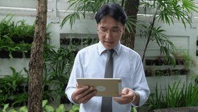 Smiling Asian businessman walking in the garden using digital tablet for webcam video call conference meeting with business team. Man using digital technology for work from home and social distancing.