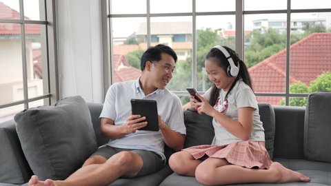Asian family Father and daughter using Smartphone and Digital tablet sitting on couch in the living room at home. lifestyle, Social Media and technology communication