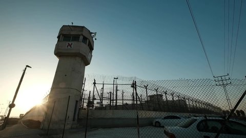 A wide angle of a prison and watchtower, last sun rays penetrating through the fence minutes before sundown and patrol vehicle is passing.