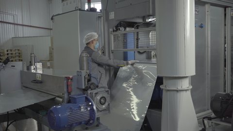 Camera approaches to serious man in face mask controlling process of manufacturing in factory. Male Caucasian employee controls processing of aluminium foil sheet by automated equipment on conveyor.
